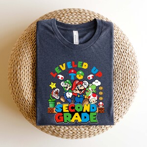 以下が含まれることがあります： スーパーマリオブラザーズのビデオゲームから、マリオとその仲間たちのグラフィックが描かれた濃い青色のTシャツ。グラフィックには「レベルアップして2年生へ」と書かれています。