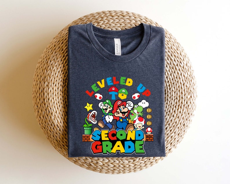 以下が含まれることがあります： スーパーマリオブラザーズのビデオゲームから、マリオとその仲間たちのグラフィックが描かれた濃い青色のTシャツ。グラフィックには「レベルアップして2年生へ」と書かれています。