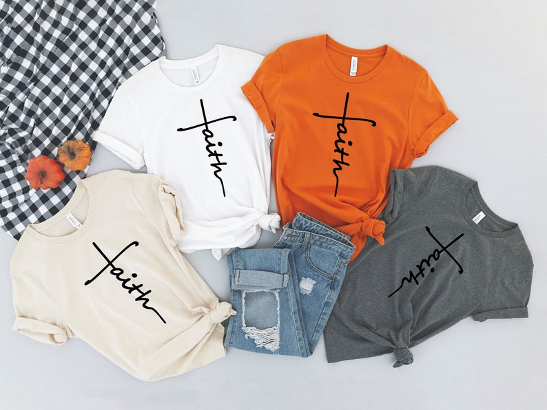 Op de afbeelding: Vier T-shirts met het woord "geloof" verticaal geschreven met een kruis voor het woord. De shirts zijn in wit, oranje, beige en grijs.