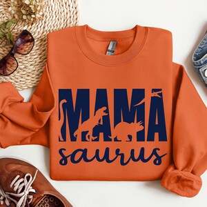May include: Orange sweatshirt with a navy blue graphic that says "Mama Saurus" with silhouettes of dinosaurs inside the letters.