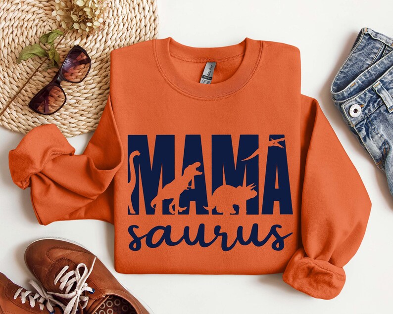 May include: Orange sweatshirt with a navy blue graphic that says "Mama Saurus" with silhouettes of dinosaurs inside the letters.