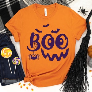 Puede incluir: Camiseta naranja con un gr&aacute;fico morado de "BOO" con una ara&ntilde;a y murci&eacute;lagos. La camiseta est&aacute; doblada y colocada sobre una superficie blanca con otras decoraciones de Halloween.