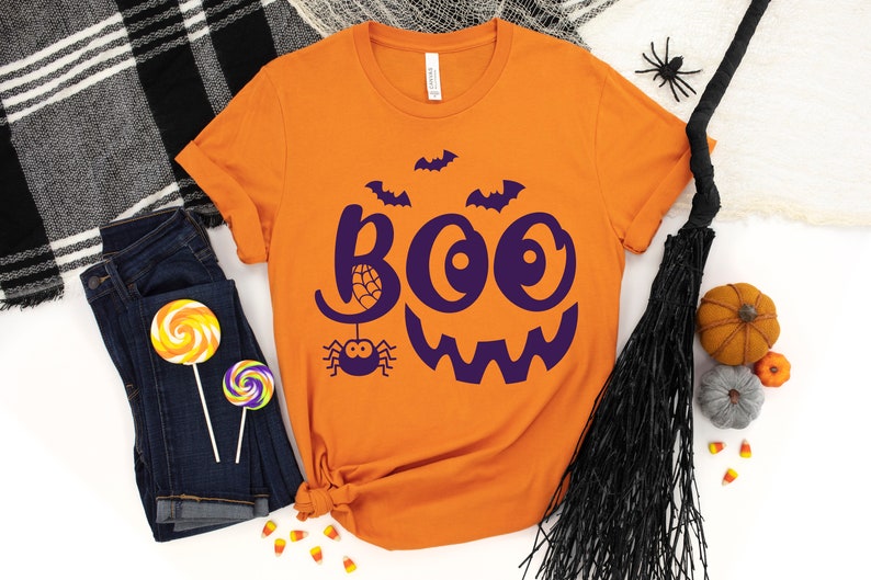 Puede incluir: Camiseta naranja con un gr&aacute;fico morado de "BOO" con una ara&ntilde;a y murci&eacute;lagos. La camiseta est&aacute; doblada y colocada sobre una superficie blanca con otras decoraciones de Halloween.