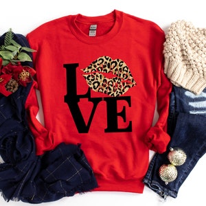 Pu&ograve; includere: Felpa rossa con una grafica nera "LOVE" e un design a labbra con stampa leopardata.