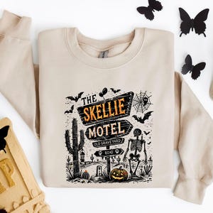 May include: Beige sweatshirt featuring a Halloween-themed graphic. The design includes a sign that reads "The Skellie Motel" with a skeleton, bats, a pumpkin, and a cactus. The sweatshirt is a neutral color, perfect for fall.