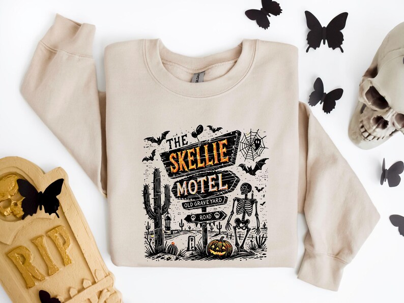 May include: Beige sweatshirt featuring a Halloween-themed graphic. The design includes a sign that reads "The Skellie Motel" with a skeleton, bats, a pumpkin, and a cactus. The sweatshirt is a neutral color, perfect for fall.