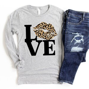 Pu&ograve; includere: Una maglietta grigia a maniche lunghe con una grafica nera "LOVE" e un design a labbra con stampa leopardata.