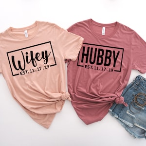 May include: Two t-shirts with the words "Wifey" and "Hubby" in black text on a white background. The shirts are pink and peach and have the date "EST. 11.17.19" printed below the words.