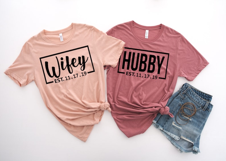 May include: Two t-shirts with the words "Wifey" and "Hubby" in black text on a white background. The shirts are pink and peach and have the date "EST. 11.17.19" printed below the words.