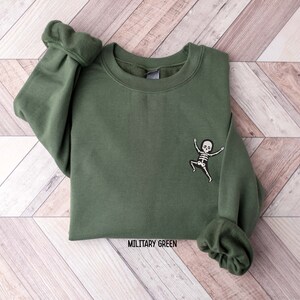 K&ouml;nnte beinhalten: Ein olivgr&uuml;ner Sweatshirt mit einem kleinen gestickten Skelett-Design auf dem linken &Auml;rmel. Der Text "MILITARY GREEN" ist unten auf dem Sweatshirt aufgedruckt.