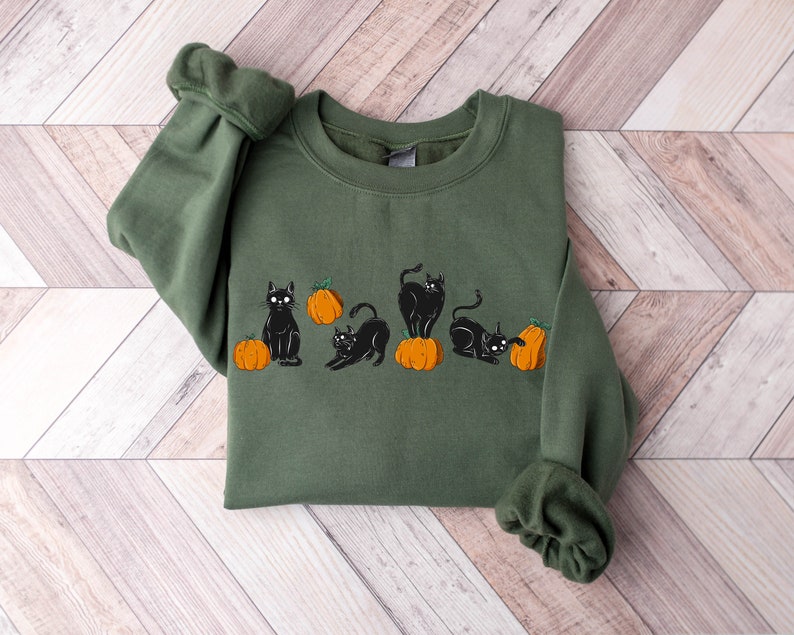 Puede incluir: Una sudadera verde con un dise&ntilde;o de gato negro y calabaza naranja. Los gatos est&aacute;n en varias poses, algunos sentados, algunos de pie y algunos mirando las calabazas.