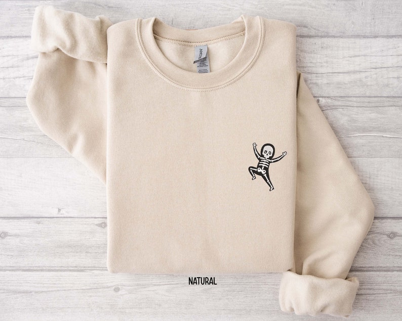 K&ouml;nnte beinhalten: Ein beiger Pullover mit einer kleinen schwarzen und wei&szlig;en Skelettgrafik eines Babyskeletts mit erhobenen Armen. Der Text "NATURAL" ist unten auf dem Pullover aufgedruckt.
