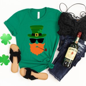 May include: Green t-shirt with a cartoon leprechaun face wearing a green hat, sunglasses, and a pipe. The shirt is paired with blue jeans, a black lace top, and black sandals.