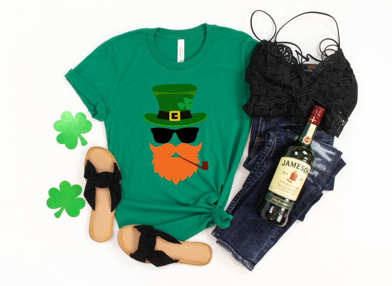 May include: Green t-shirt with a cartoon leprechaun face wearing a green hat, sunglasses, and a pipe. The shirt is paired with blue jeans, a black lace top, and black sandals.