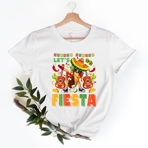 May include: White t-shirt with a cartoon illustration of a taco wearing a sombrero and sunglasses, holding a guitar and doing the dab. The text "Let's Fiesta" is printed below the illustration.