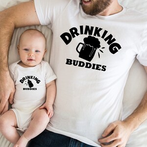 May include: A white t-shirt with the text "Drinking Buddies" and a beer mug graphic. A white onesie with the text "Drinking Buddies" and a baby bottle graphic. A father and baby are wearing the shirts.