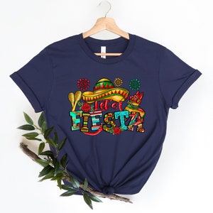 Puede incluir: Camiseta azul marino con un dise&ntilde;o gr&aacute;fico colorido que incluye un sombrero, maracas y el texto "Let's Fiesta".