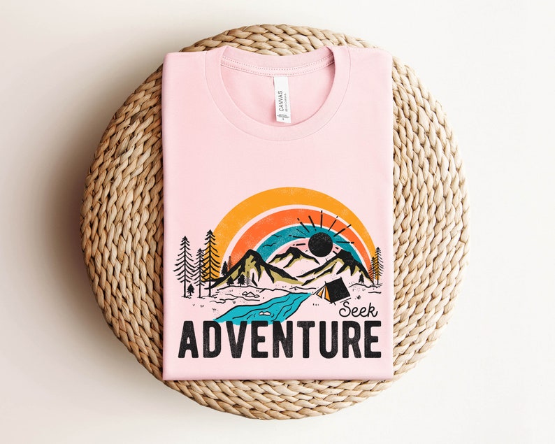 May include: A light pink t-shirt with a colorful graphic design. The design features a rainbow, mountains, trees, a river, and a tent. The words "Seek Adventure" are printed in black block letters below the graphic.