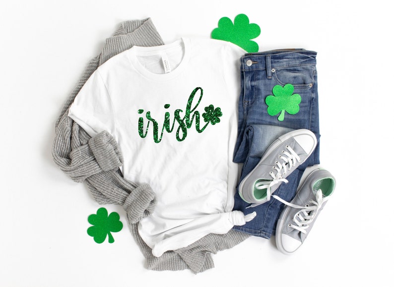 May include: White t-shirt with the word "irish" in green glitter script and a green glitter shamrock. The shirt is paired with blue jeans, gray sneakers, and a gray sweater. Green shamrock cutouts are also included.