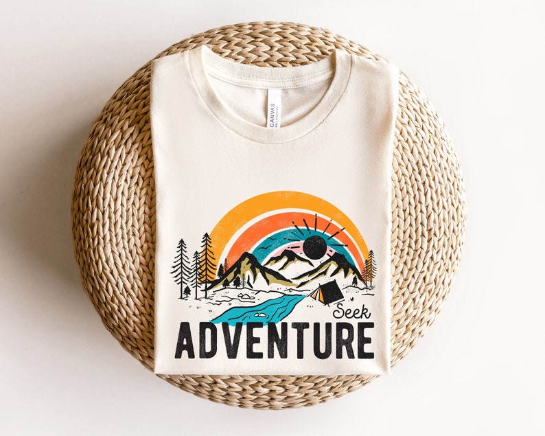 May include: Cream-colored t-shirt featuring a colorful graphic of a mountain landscape with a rainbow, sun, and river. The text "Seek Adventure" is printed in bold, black letters below the image. The shirt is made of a soft material.
