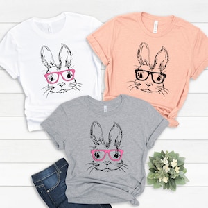 May include: Three t-shirts with a black and white illustration of a bunny wearing pink glasses. The shirts are in white, peach, and heather grey.