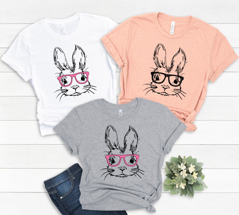 May include: Three t-shirts with a black and white illustration of a bunny wearing pink glasses. The shirts are in white, peach, and heather grey.
