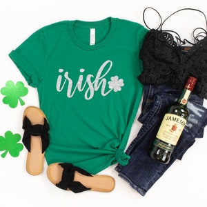 May include: A green t-shirt with the word "irish" in silver glitter script and a glitter shamrock. The image also includes black sandals, a black lace top, dark wash jeans, and a bottle of Jameson Irish Whiskey.