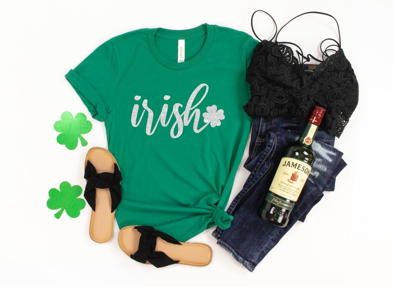 May include: A green t-shirt with the word "irish" in silver glitter script and a glitter shamrock. The image also includes black sandals, a black lace top, dark wash jeans, and a bottle of Jameson Irish Whiskey.