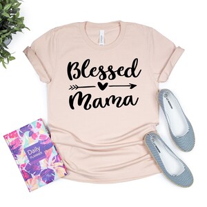 May include: A light pink t-shirt with the text "Blessed Mama" in black lettering with an arrow and heart graphic.