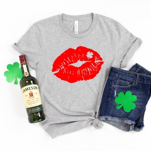 May include: A gray t-shirt with a red lipstick print and a shamrock. The shirt is paired with denim shorts and a bottle of Jameson Irish Whiskey. There are green shamrocks on the shirt and shorts.