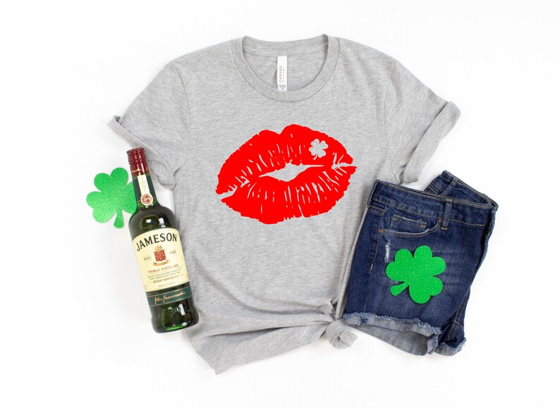 May include: A gray t-shirt with a red lipstick print and a shamrock. The shirt is paired with denim shorts and a bottle of Jameson Irish Whiskey. There are green shamrocks on the shirt and shorts.
