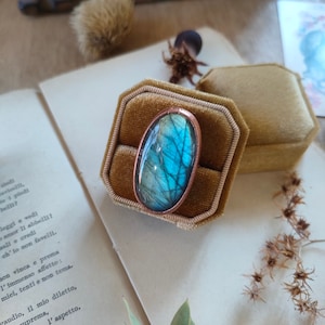 Könnte beinhalten: Ein Ring mit einem großen ovalen blauen Labradorit-Edelstein, der in Kupfer gefasst ist. Der Ring wird in einer Samt-Ringbox präsentiert.