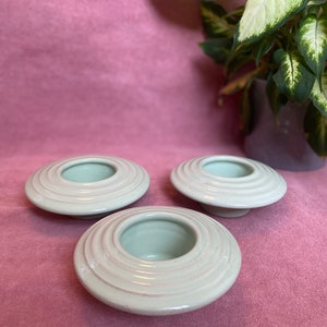 May include: Three white ceramic candle holders with a ringed design. The holders are on a pink surface.