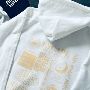 Puede incluir: Sudadera con capucha blanca con un estampado dorado que dice "Escape the Ordinary" e incluye frases como "Breathe Repeat", "See the Good in Small Things", "Every Moment Matters" y "Simplicity is Courage Over Comfort".