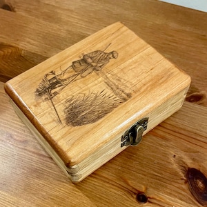 Pode incluir: Uma caixa de madeira com tampa articulada e fecho de metal. A parte superior da caixa apresenta uma gravura de uma pessoa num campo com uma espingarda. A caixa é feita de madeira clara.