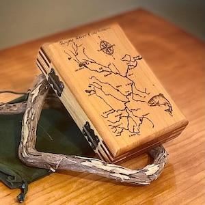 May include: A wooden box with a hinged lid, engraved with a map of the Boyne River and a compass. The box is made of light wood with dark wood accents. The lid is open and the inside of the box is visible.
