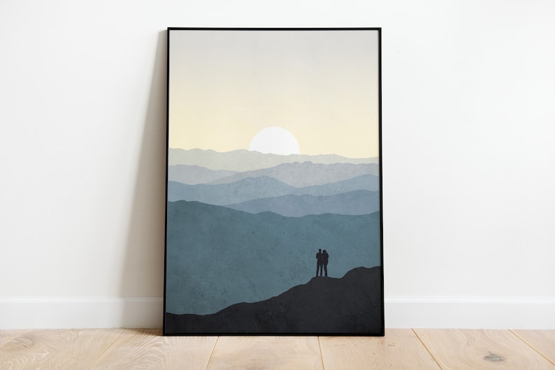 May include: A framed print of a minimalist landscape with a couple standing on a mountaintop, silhouetted against a pale yellow sky with a white sun. The mountains are layered in shades of blue and gray.