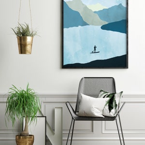 May include: A framed art print depicts a serene landscape with a person paddleboarding on a lake, mountains, and a large sun. A black metal chair with pillows, a potted plant, and a hanging plant in a gold pot complete the interior scene.