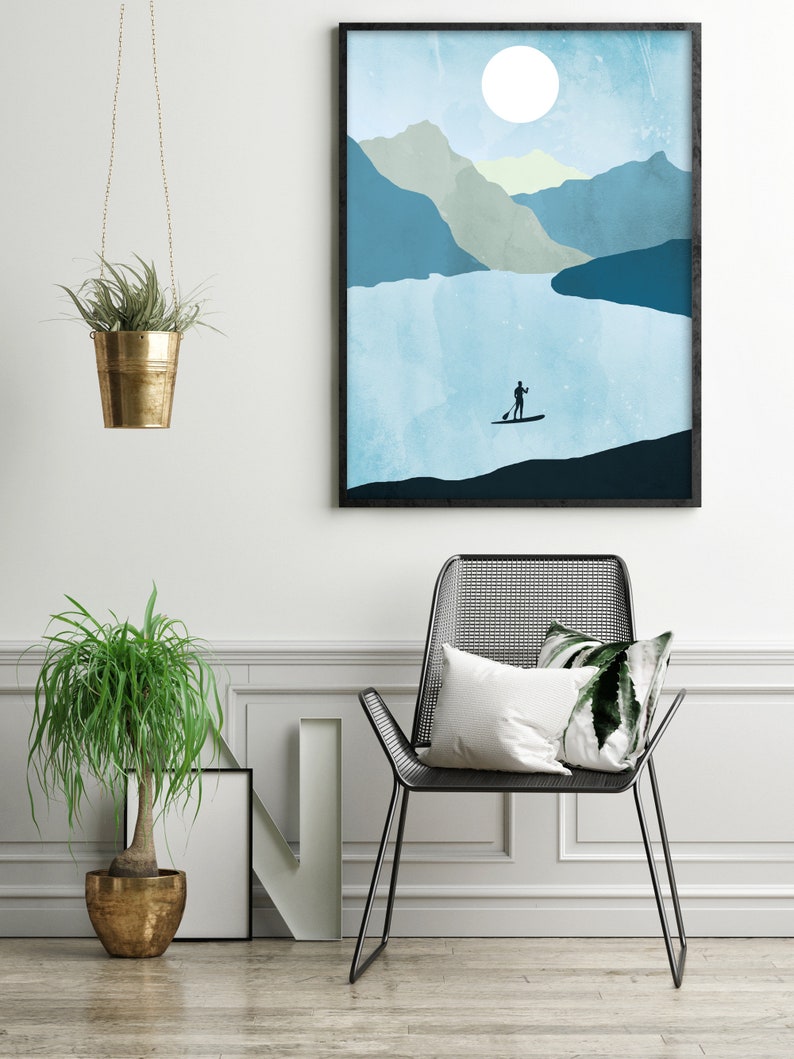May include: A framed art print depicts a serene landscape with a person paddleboarding on a lake, mountains, and a large sun. A black metal chair with pillows, a potted plant, and a hanging plant in a gold pot complete the interior scene.
