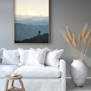 May include: A framed print of a minimalist landscape with a couple silhouetted against a mountain range and a sun setting behind them. The print is in shades of blue and yellow and is hanging on a gray wall.