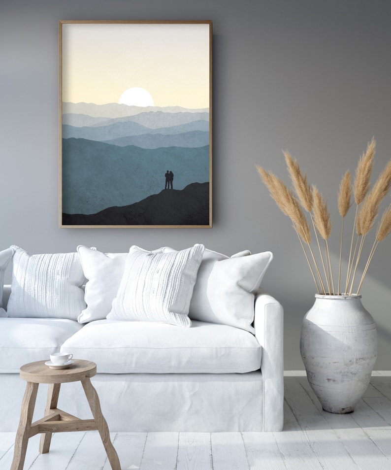May include: A framed print of a minimalist landscape with a couple silhouetted against a mountain range and a sun setting behind them. The print is in shades of blue and yellow and is hanging on a gray wall.