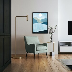 May include: An interior shot featuring a framed landscape art print with a person paddleboarding on a lake. A light green armchair sits in front of the artwork. A gold floor lamp and a side table with a vase of flowers are also visible.