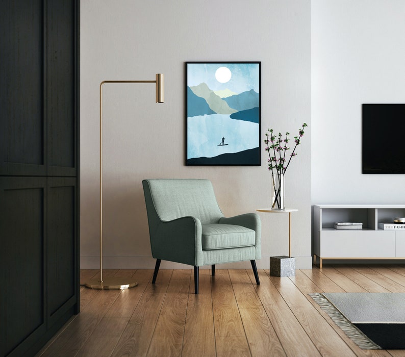 May include: An interior shot featuring a framed landscape art print with a person paddleboarding on a lake. A light green armchair sits in front of the artwork. A gold floor lamp and a side table with a vase of flowers are also visible.