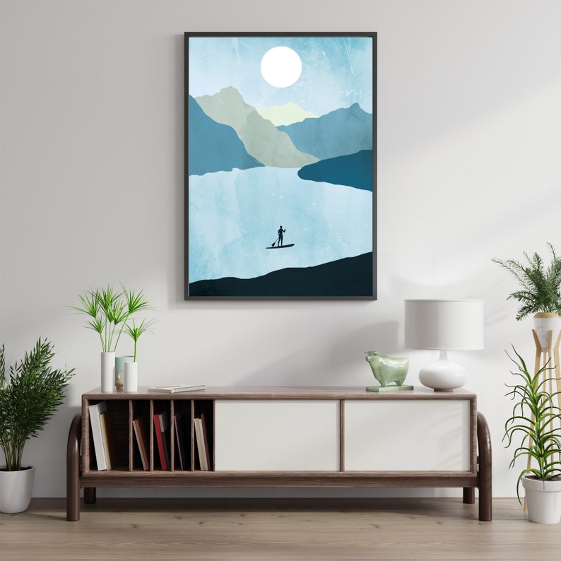 May include: Abstract art print featuring a serene mountain lake scene. A silhouetted figure paddles a stand-up paddleboard on the water under a large white moon. The artwork is framed in a black border and evokes a sense of tranquility.