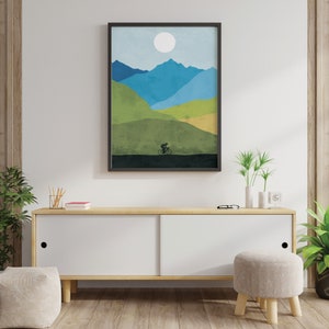 May include: A framed abstract landscape print with a silhouette of a person riding a bicycle in front of a mountain range with a white sun in the sky. The print is in shades of blue, green, and brown.