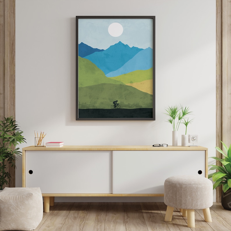 May include: A framed abstract landscape print with a silhouette of a person riding a bicycle in front of a mountain range with a white sun in the sky. The print is in shades of blue, green, and brown.