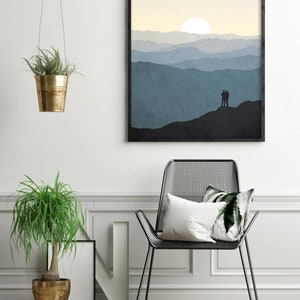 May include: A framed print of a minimalist landscape with a couple silhouetted against a mountain range and a sun setting in the distance. The print is hanging on a white wall above a black metal chair with a white pillow and a green pillow.
