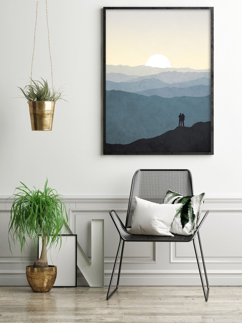 May include: A framed print of a minimalist landscape with a couple silhouetted against a mountain range and a sun setting in the distance. The print is hanging on a white wall above a black metal chair with a white pillow and a green pillow.