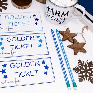 以下が含まれることがあります： 白い木製の表面に、青い星と「GOLDEN TICKET redeem for... 」という文字が書かれた、白い印刷可能な金のチケットカードが3枚。茶色の木製の星と雪の結晶が置かれている。