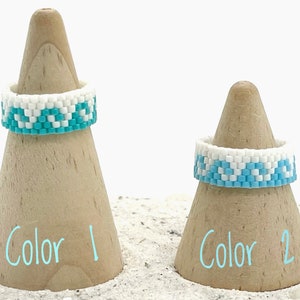 May include: Two beaded rings on wooden ring displays. The rings are made of white and turquoise beads in a geometric pattern. The display on the left is labeled "Color 1" and the display on the right is labeled "Color 2".